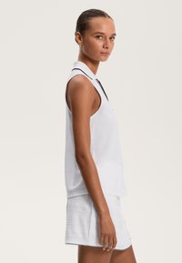 Kobieta w białej bezrękawnikowej bluzce z kołnierzykiem i białych szortach o teksturze stoi bokiem na jednolitym jasnym tle.