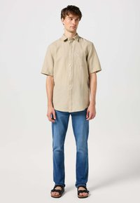 Camicia beige a maniche corte con bottoni e taschino sul petto, abbinata a jeans blu e sandali neri. Tessuto liscio, vestibilità comoda, design minimalista.