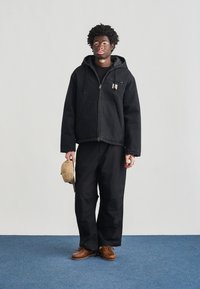 Μαύρο τζιν μπουφάν με κουκούλα, μπροστινό φερμουάρ και τσέπη στο στήθος. Συνδυασμένο με μαύρο παντελόνι εργασίας και καφέ παπούτσια, κρατώντας ένα μπεζ καπέλο.