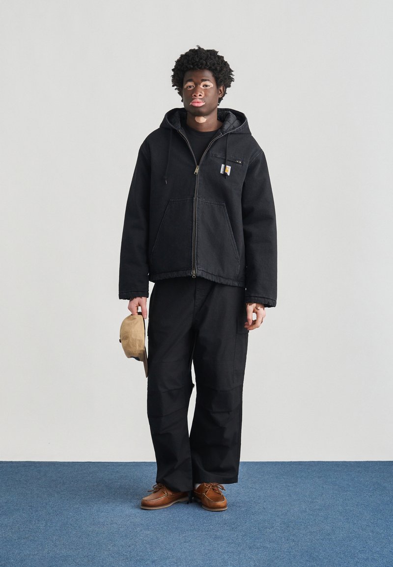 Veste en jean noire avec capuche, fermeture éclair frontale et poche poitrine. Portée avec un pantalon de travail noir et des chaussures marron, tenant un chapeau beige.