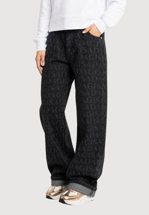 Personne portant un jean noir avec le motif répété "SADAGIS", un sweat-shirt blanc et des baskets épaisses métalliques couleur or rose, debout avec une main dans la poche.
