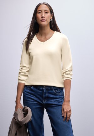 Crèmekleurige pullover met V-hals en lange mouwen, gecombineerd met blauwe denim jeans. De stof is soepel met een lichte rek. Het model houdt een grijze jas vast.