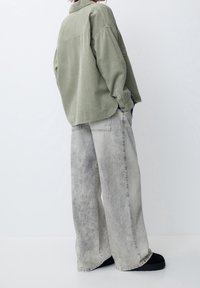 Camisa de corduroy verde oversized con una superficie texturizada, combinada con vaqueros de pierna ancha gris claro que presentan un patrón desvanecido y bolsillos laterales.