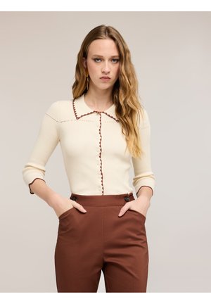 Femme aux longs cheveux ondulés portant un pull crème boutonné avec bordure marron et un pantalon marron taille haute, mains dans les poches, fond neutre.