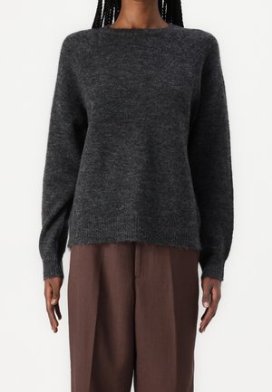 Personne portant un pull en tricot duveteux gris foncé à manches longues et un pantalon marron à jambes larges sur un fond blanc uni.