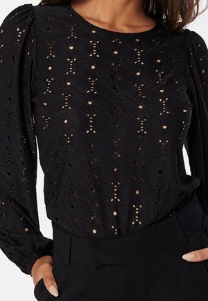 Blouse noire à manches longues avec motif en dentelle ajourée florale et épaules bouffantes, associée à un pantalon noir taille haute.