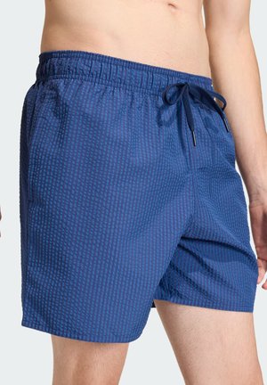 Man draagt blauwe zwemshorts met patroon, elastische tailleband en trekkoord, staand tegen een effen lichte achtergrond.