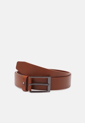 Ceinture en cuir marron roulée avec boucle métallique rectangulaire et petite étiquette rouge, blanche et bleue près de la boucle sur un fond uni.