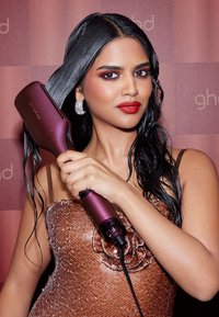 Outil de coiffage GHD de couleur violet foncé, de forme rectangulaire, avec une finition brillante. Le modèle tient l'appareil avec des cheveux noirs mouillés et lisses, portant un haut en sequins.