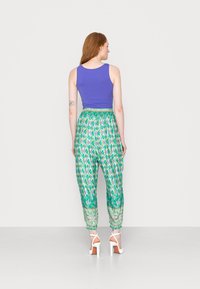 Top roxo sem mangas combinado com calças harem verdes estampadas que apresentam uma cintura elástica e punhos, complementado por sandálias de salto brancas.