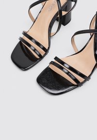 Sandalias de tacón negro con punta cuadrada, acabado de charol y tres tiras finas a través del pie para mayor soporte. Plantilla neutral con ligera textura.