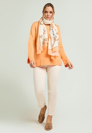 Frau trägt ein pfirsichfarbenes Langarmshirt, hellbeige Hose, braune Tassel-Loafers und einen weißen Schal mit Kettenmuster, geht auf die Kamera zu.