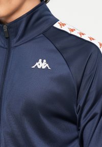 Mornariće plava atletska jakna s visokim ovratnikom, zatvaračem na zip, i bijelom akcentom s ponovljenim crvenim logotipom na ramenu. Glatka tekstura.