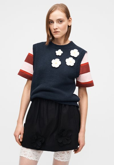 Marineblauer Strickpullover mit drei weißen Blumen-Applikationen über einem gestreiften Kurzarmhemd, kombiniert mit einem schwarzen Rock mit Blumenakzenten.