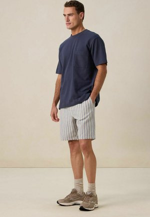 Marineblauw T-shirt met korte mouwen en zak, gecombineerd met meerkleurige gestreepte shorts. Model draagt beige sneakers en lichtgekleurde sokken.