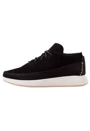 Black suede low-top sneaker with black laces, white thick sole, and white stitched detail on the heel, shown from the side on a white background.
