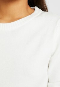 Vit, mjuk stickad tröja med ribbad rund halsringning, som visar en jämn yta och en ren, minimalistisk design. Inga mönster eller detaljer synliga.