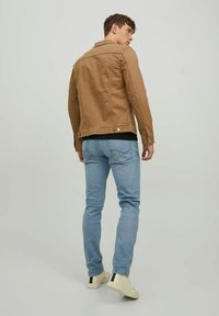 Chaqueta de algodón color marrón claro con un diseño ajustado, botones a presión y detalle de yugo en la parte trasera, combinada con jeans azul claro y zapatillas beige.