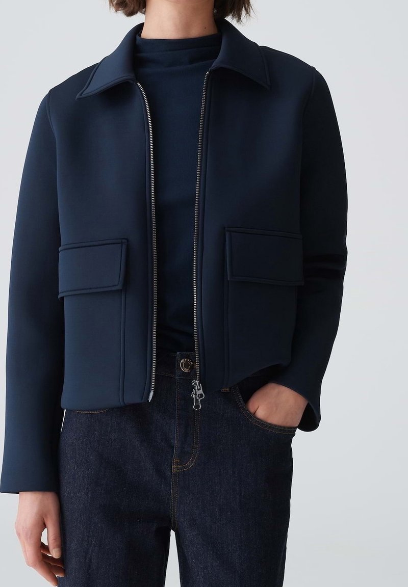 Giacca corta blu navy in tessuto liscio, con cerniera frontale, due tasche con pattina e colletto appuntito, abbinata a jeans scuri.