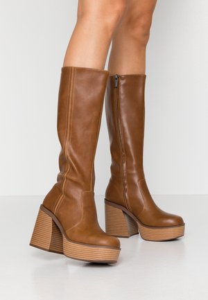 Knee-high brown leather platform boots with block heels worn on bare legs, featuring side zippers and stacked wooden soles.