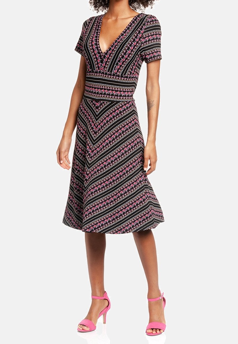 Femme portant une robe noire avec des motifs diagonaux roses et blancs et des sandales à talons roses, debout sur un fond blanc.