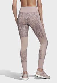 Žena v pásových světle purpurových legínách s hadím vzorem, doplněných o odpovídající sportovní boty, zobrazena zezadu.