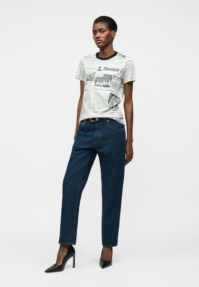 Γυναίκα με μαύρα τακούνια, σκούρο μπλε τζιν και λευκή μπλούζα Moschino με τύπωμα εφημερίδας, μπροστά σε απλό γκρι φόντο.