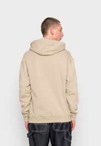 Beige hoodie with a spacious hood, ribbed cuffs, and hem. Soft texture visible; worn with dark denim pants. Back view shows seam details.