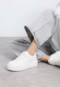 Zapatillas de plataforma blancas con paneles texturizados, perforaciones y un acento naranja en la lengüeta. Combinadas con jeans de pierna ancha gris claro.