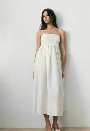 Naine valges varrukateta midi kleidis, õhukeste rihmadega, paljajalu seisab ühevärvse heleda seina ääres, vaadates veidi alla ja küljele.