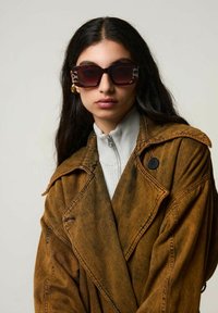 Bruine oversized denimjack met grote revers, grijze trui met ritssluiting eronder en donkere zonnebril met dierenprintaccenten op de zijkanten.