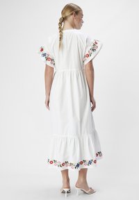 Abito di cotone bianco con maniche corte a campana, caratterizzato da un vivace ricamo floreale lungo le maniche e l'orlo. Design fluido e parte inferiore arricciata.