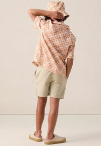 Korte mouwen, perzikpatroon shirt, lichtbeige shorts en nude sneakers. Het shirt heeft een herhalend ontwerp van zon en planten.