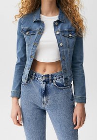 Giacca di jeans in azzurro chiaro con un design corto, dotata di due tasche sul petto, bottoni argentati e colletto classico. Indossata sopra una maglietta bianca.