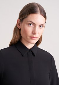 Camicia nera con colletto strutturato e texture liscia, caratterizzata da una patta nascosta e un design minimalista.