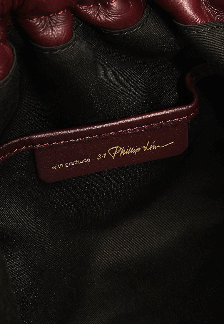 Bordowa skórzana metka z złotym napisem "z wdzięcznością" oraz "3:1 Phillip Lim", przymocowana do czarnej tkaniny wewnętrznej.