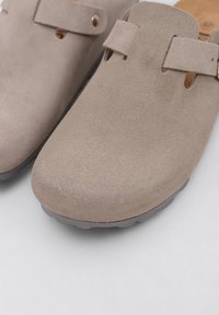 Beige suede clogs with rounded toe, two strap openings, and light gray rubber sole. Features a smooth texture and minimalistic design.