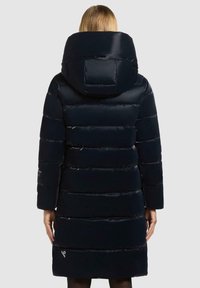 Parka blu navy imbottita con un grande cappuccio, caratterizzata da sezioni trapuntate orizzontali e una superficie liscia e lucida. Presenta dettagli con logo sulla manica.