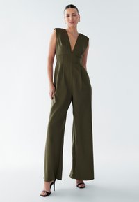 Un combine-pantalon vert olive avec un décolleté en V profond, des manches courtes, un corsage ajusté et un pantalon évasé, complété par des sandales à talons noires.