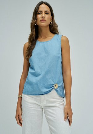 Mujer con cabello largo, con pendientes de aro dorados, top sin mangas azul claro con detalle de botones y pantalones blancos sobre fondo liso.