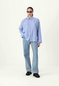 Camisa de botão às riscas azul e branca, de corte descontraído, combinada com jeans flare azul claro e sapatos pretos, usada com óculos de sol.