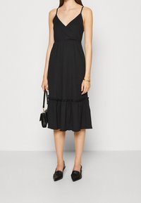 Robe noire midi avec bretelles fines, décolleté en V, corsage ajusté et ourlet froncé. Portée avec des mules noires et tenant un petit sac à main.