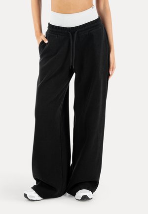 Kobieta w szerokich czarnych spodniach dresowych z wiązanym w pasie sznurkiem i białych tenisówkach, stojąca z jedną ręką w kieszeni.