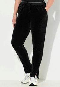 Zwarte fluwelen joggingbroek met een trekkoord in de taille, zijzakken en splitten bij de enkels, gecombineerd met witte sportschoenen.