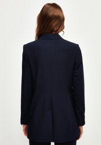 Blazer blu navy con vestibilità sartoriale, colletto a revers e bottoni decorativi sulle maniche. Tessuto liscio e design strutturato con cuciture visibili.
