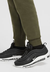 Black Nike Air Max 97 shoes with wavy texture and white accents, paired with olive green sweatpants featuring ribbed cuffs.