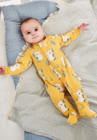 Gul babydragt med koala- og bladmotiv, lange ærmer, fuld lynlås foran, blødt stof, ligger på en tekstureret grå tæppe og pude.