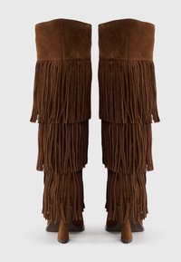 Botas de ante marrón hasta la rodilla con flecos largos a los lados y un tacón estilizado. Textura suave con la parte superior ligeramente redondeada.