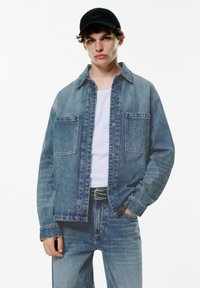 Lichtblauwe denimjack met twee voorzakken, knoopsluiting en een losse pasvorm, gecombineerd met een wit shirt en blauwe jeans. Zwarte pet gedragen.