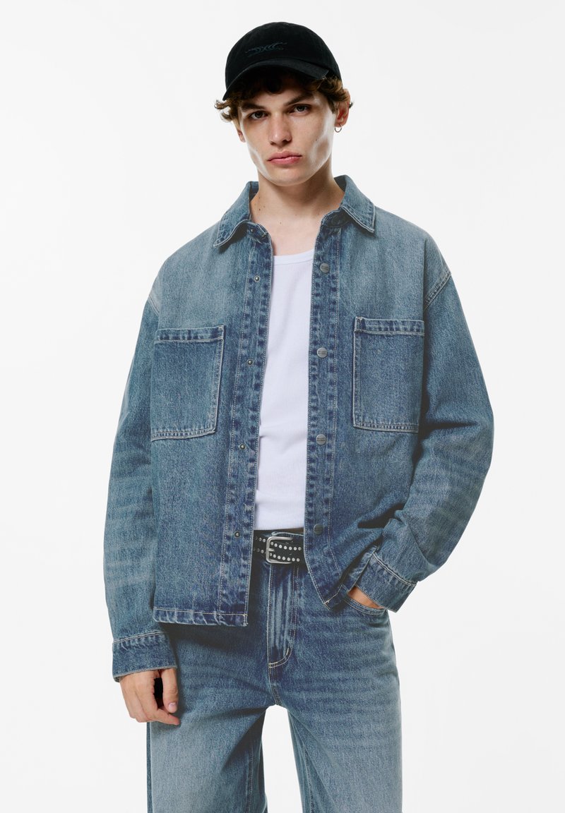Giacca di jeans azzurro chiaro con due tasche frontali, chiusura con bottoni e vestibilità ampia, abbinata a una camicia bianca e jeans blu. Cappellino nero indossato.
